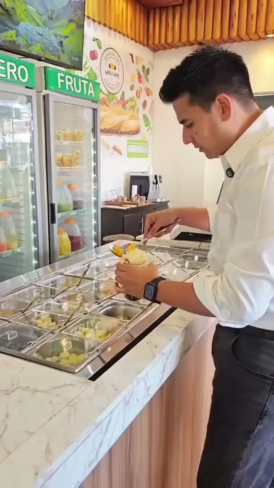 Un cliente arma su vaso en el mostrador de Quiero Fruta, eligiendo fruta, toppings y salsas, y después lo disfruta en la mesa