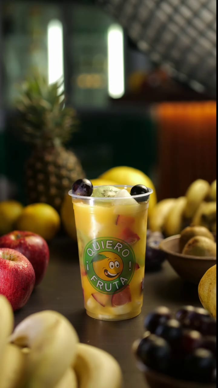 Un vaso de ensalada de frutas con uvas, rodeado de ananá, manzanas y bananas